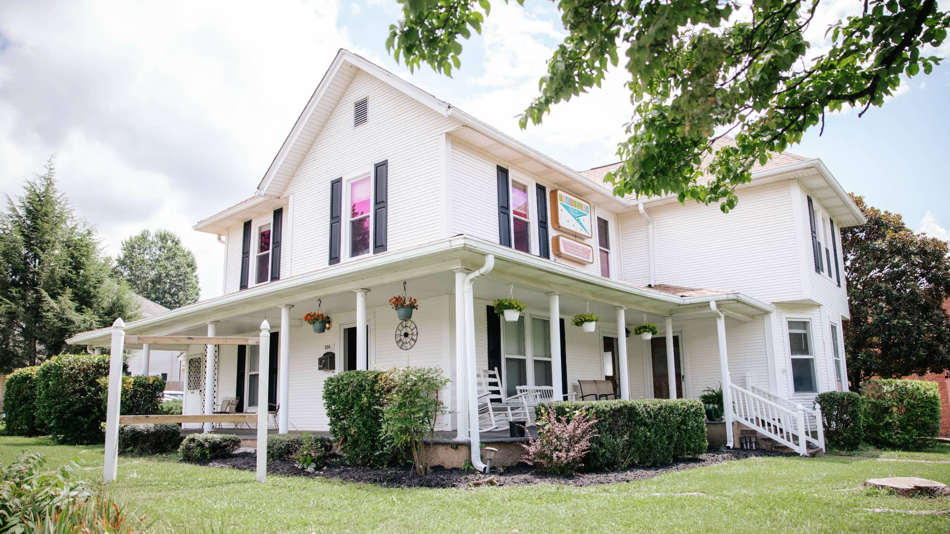 A white two-story house with black shutters and colorful flower pots on the porch, surrounded by a green lawn.