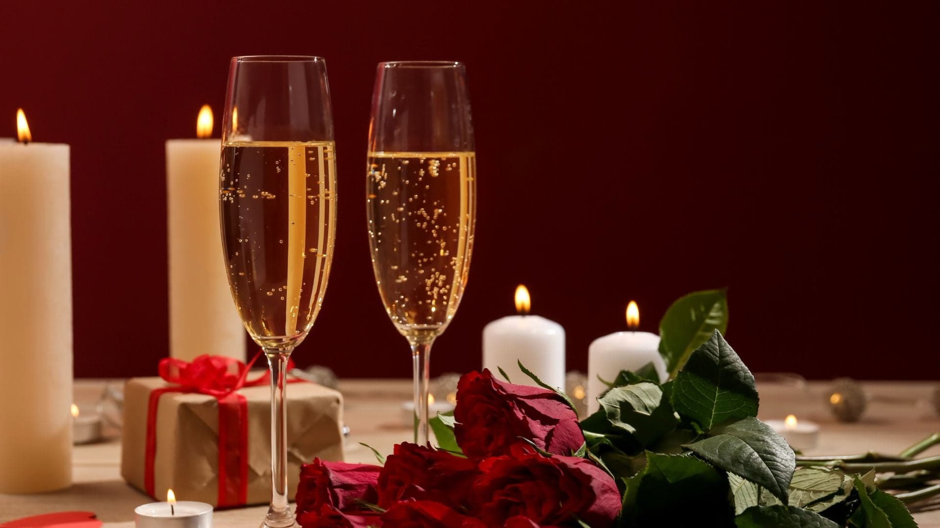Two champagne flutes beside red roses, candles, and a gift box against a deep red background.