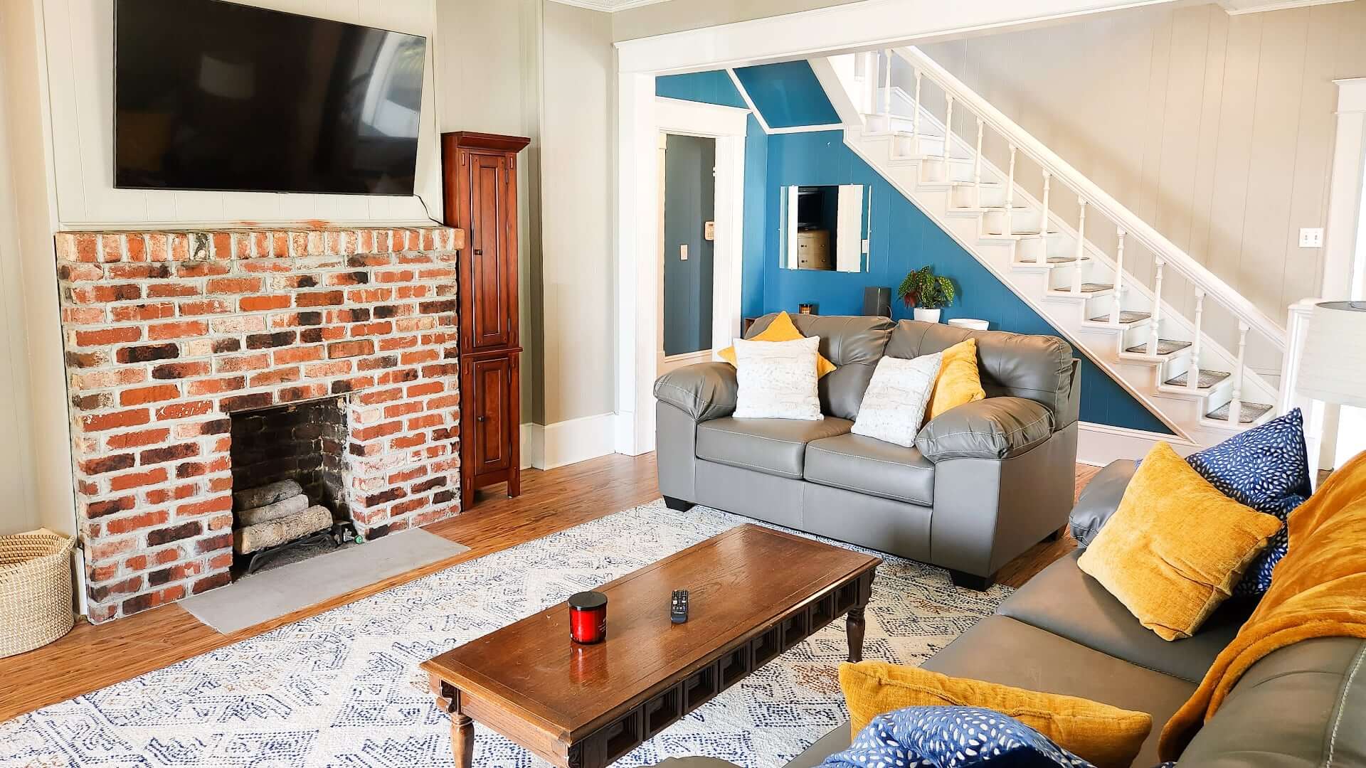 A cozy living room with a brick fireplace, gray sofas, and a staircase leading to a blue-walled area.